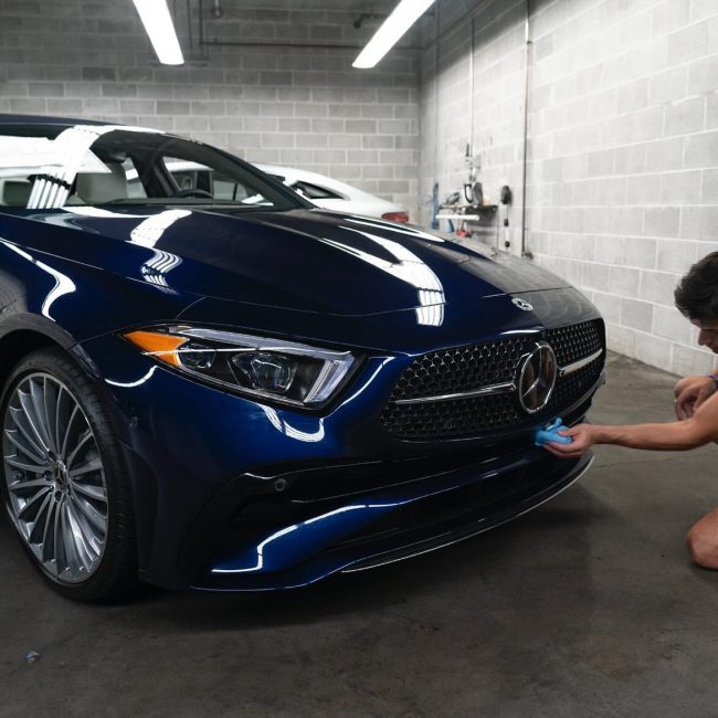 Detailer applying ceramic coating to the front bumper of a dark blue Mercedes-Benz in a professional studio setting