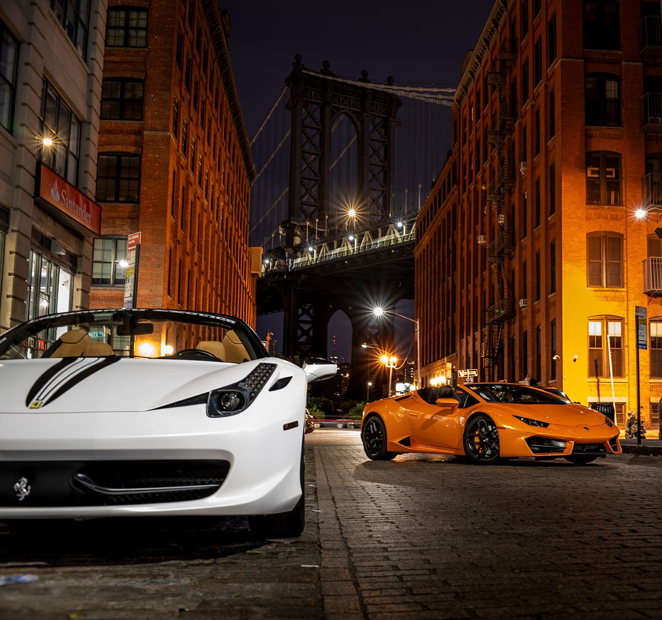White Ferrari and orange Lamborghini parked on cobblestone street in NYC at night with Manhattan Bridge in the background, showcasing high-gloss paint protection film