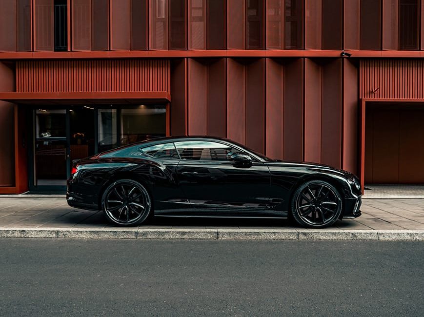 Glossy black luxury coupe parked in front of modern red building after paint correction and PPF application