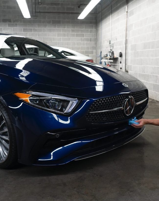 Detailer applying ceramic coating to the front bumper of a dark blue Mercedes-Benz in a professional studio setting
